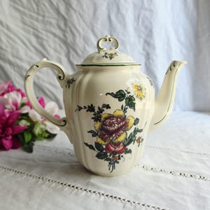 May include: A cream-colored ceramic teapot featuring a fluted body and a lid with a decorative handle. The surface is adorned with a colorful floral pattern of yellow, red, and purple blossoms with green leaves and thin green trim accents.