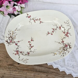 May include: A large, oval, white serving platter with a delicate floral design. The design features pink and white flowers with brown stems and leaves.