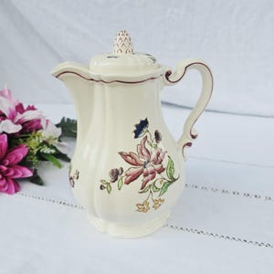 May include: Cream-colored ceramic pitcher with a lid, featuring a floral design in shades of pink, blue, and green. The pitcher has a curved handle and a spout. The rim and handle are accented with a dark red trim. A decorative piece for serving beverages.
