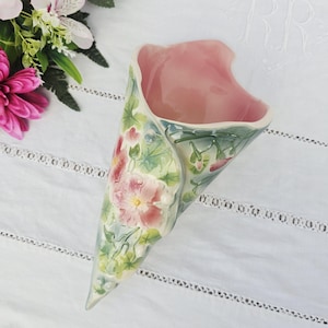 May include: A ceramic wall pocket vase in a cone shape, featuring a pink interior and a floral design on the exterior. The vase has a light green and pink color scheme with detailed flower and leaf patterns.