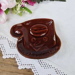 Peut inclure: Un plat en céramique marron en forme de tasse à café avec un motif de grain de café et un visage souriant. Le plat a une anse et une base de soucoupe. Le texte "Cafés Rosa Camara" est écrit sur la soucoupe.