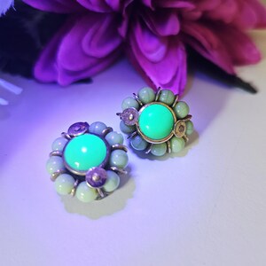 Pode incluir: Um par de brincos redondos com um cabochão central verde néon brilhante rodeado por um anel de contas verde-pálido e pequenos detalhes metálicos. Os brincos exibem um brilho vibrante sob luz ultravioleta, destacando o contraste entre o verde néon central e as contas verde-pálido circundantes.