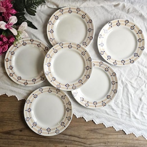 Peut inclure: Ensemble de six assiettes en céramique blanche. Chaque assiette présente une bordure décorative avec un motif floral bleu répété et de fines rayures orange et marron. Les assiettes sont disposées sur un tissu blanc.