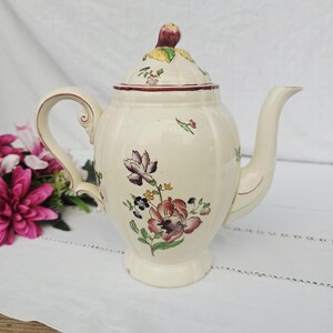 May include: Cream-colored ceramic teapot with a floral design in shades of red, purple, and green. The teapot has a curved handle, a spout, and a lid with a decorative knob. The floral pattern is hand-painted, adding to its vintage charm.