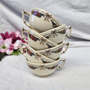 Puede incluir: Una pila de cinco tazas de té color crema. Cada taza presenta un diseño decorativo floral y de lazo en rojo, azul y amarillo. Las tazas tienen un diseño de asa clásico y están apiladas, mostrando su estética vintage.