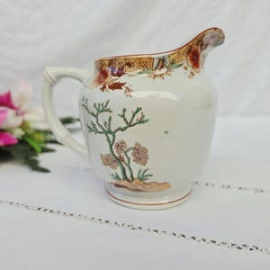 Peut inclure: Un pot à lait en céramique de couleur crème orné d'un motif peint à la main représentant un arbre stylisé vert avec un feuillage brun et des accents floraux orange près du bord. Le pot possède une anse courbée et un large bec verseur.