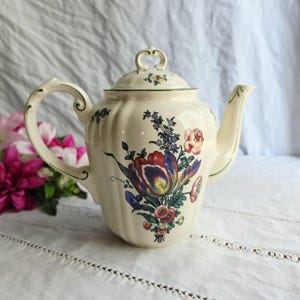 May include: A cream-colored ceramic teapot with a fluted body and a matching lid. The teapot features a colorful floral bouquet illustration with red, purple, and blue flowers, accented by green stems and leaves, and a curved handle and spout.