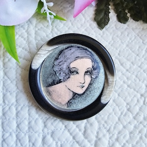 Peut inclure: Broche ronde noire et blanche représentant une illustration de style Art Déco d'un visage de femme. La femme a les cheveux gris et se détache sur un fond bleu clair. La broche a une finition brillante.