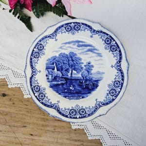 Może przedstawiać: Niebieski i biały talerz ceramiczny z ozdobną obwódką i sceną przedstawiającą chatkę nad rzeką. Na talerzu nadrukowany jest tekst "The Hay-Wain by Constable".