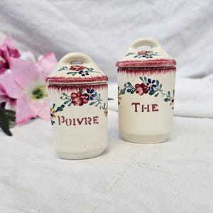 Peut inclure: Deux pots à épices en céramique blanc cassé avec couvercles. Les couvercles et les bords sont décorés d'une bordure bordeaux et de motifs floraux. Un pot porte l'inscription "POIVRE" en bordeaux, l'autre "THE".