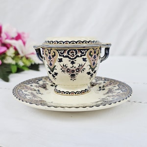 Pode incluir: Um conjunto de chávena de caldo e pires em cerâmica cor de creme com um padrão decorativo em azul escuro, rosa e preto. A chávena tem duas pequenas pegas nos lados opostos e ambas as peças estão adornadas com motivos florais e volutas.