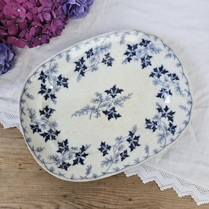 Könnte beinhalten: Eine ovale, antike Servierplatte mit cremefarbenem Grund und blauem Blumendesign. Das blaue Blumenmuster umrandet den Rand und die Mitte der Platte. Die Platte liegt auf einem weißen Tuch mit Spitzenbesatz.
