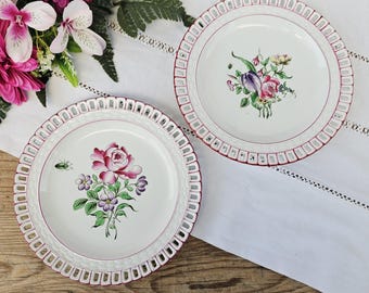 May include: Two white ceramic plates with floral designs. Each plate has a decorative border with a pink trim. One plate features a pink rose and purple flowers, while the other has a bouquet of flowers including a tulip. The plates are on a white tablecloth.