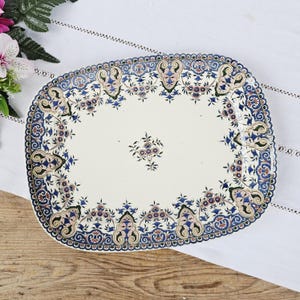 Peut inclure: Un plat de service ovale en céramique avec un centre de couleur crème orné d'un petit motif floral. La bordure est décorée d'un motif complexe de volutes, d'éléments floraux et de formes géométriques symétriques en bleu, vert et brun.
