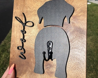 May include: Wooden sign with a black silhouette of a dog's rear end with a hook attached. The sign says "Woof" in a cursive font.