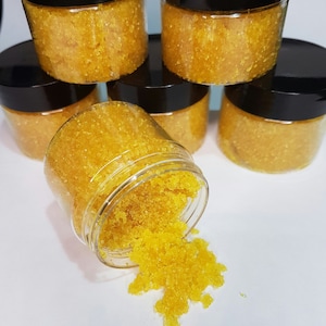 May include: Several small glass jars filled with a yellow sugar scrub. The jars have black lids and are arranged in a pyramid shape. One jar is tipped over, spilling the scrub onto a white surface.