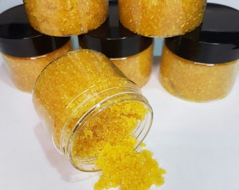 May include: Multiple small glass jars filled with a yellow sugar scrub. The jars have black lids and are arranged in a pyramid shape. One jar is tipped over, spilling the scrub onto a white surface.