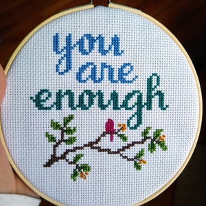 May include: A white embroidery hoop with a cross-stitch design that reads "you are enough" in blue and green thread. A small red bird is perched on a brown branch with green leaves and orange flowers.