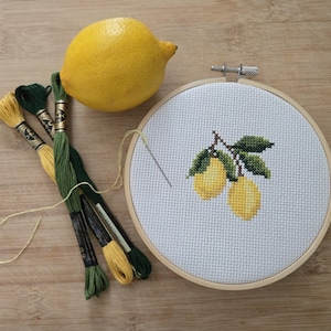 Puede incluir: Un bastidor de bordado de punto de cruz con un diseño de limón, junto con un limón real y carretes de hilo de bordar verde y amarillo. El bastidor es redondo y de madera.