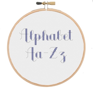 May include: A close-up of a wooden embroidery hoop with white Aida cloth stretched across it. The cloth has a cross-stitch design of the alphabet in a blue-grey color. The text reads "Alphabet A-Z".