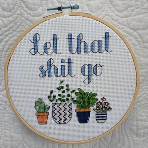 Peut inclure: Un cercle de broderie en bois rond avec un tissu blanc tendu dessus. Le tissu est brodé en bleu et vert et porte l'inscription "Let that shit go" et représente quatre plantes en pot.