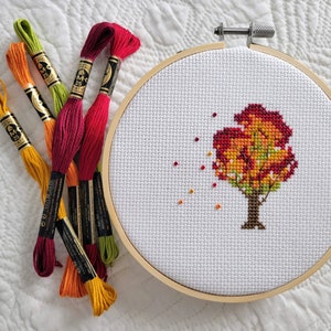 May include: A close-up of a cross-stitch embroidery hoop with a colorful autumn tree design. The hoop is surrounded by several spools of embroidery floss in various shades of red, orange, yellow, and green.