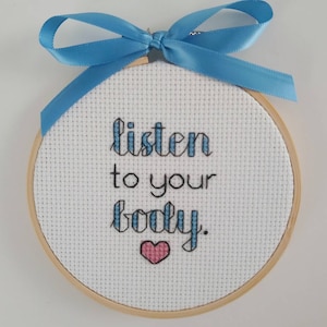 Op de afbeelding: Een witte borduurring met een blauw lint eromheen gebonden. De ring heeft een kruissteekontwerp dat "luister naar je lichaam" zegt met een roze hart onder de tekst.