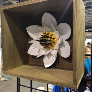 May include: A white ceramic flower sculpture with yellow and black accents is displayed in a wooden frame. The flower has a geometric design and is a unique piece of art.