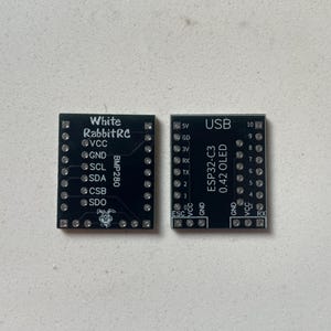 Könnte beinhalten: Zwei schwarze rechteckige elektronische Leiterplatten mit weißem Aufdruck und Stiftlöchern. Die linke Platine ist mit White RabbitRC und BMP280 beschriftet. Die rechte Platine trägt die Aufschrift USB, ESP32-C3 und 0.42 OLED mit Pin-Bezeichnungen.