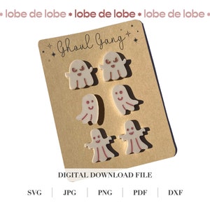 May include: A set of six white ghost-shaped earrings with pink facial features. The earrings are displayed on a brown card with the text "Ghoul Gang" in black cursive lettering.