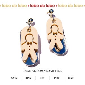 Puede incluir: Un par de pendientes colgantes con un diseño de hombre de jengibre. Los pendientes tienen un recorte de hombre de jengibre de color beige claro sobre un respaldo espejado azul y plateado. Los pendientes están suspendidos de un poste plateado.