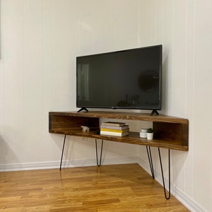 May include: A dark brown, corner TV stand with black hairpin legs. The stand has an open shelf with books, small plants, and a decorative bear figurine. A black flat-screen TV sits on top. The stand is against a white wall.