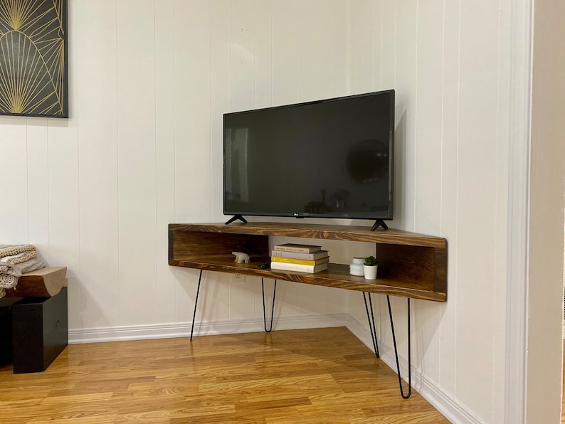 May include: A dark brown, corner TV stand with black hairpin legs. The stand has an open shelf with books, small plants, and a decorative bear figurine. A black flat-screen TV sits on top. The stand is against a white wall.