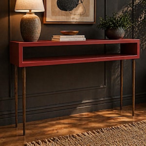 May include: A red console table with a rectangular design and brass-colored legs. The table has a shelf and is placed against a dark wall. A lamp, books, and a bowl are on the table. The table is approximately 30 inches long.