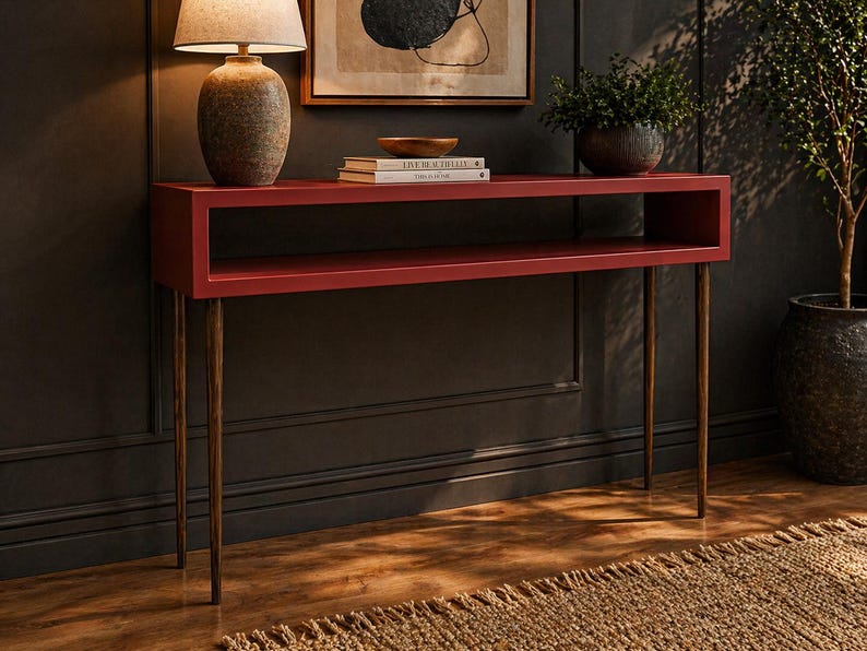 May include: A red console table with a rectangular design and brass-colored legs. The table has a shelf and is placed against a dark wall. A lamp, books, and a bowl are on the table. The table is approximately 30 inches long.