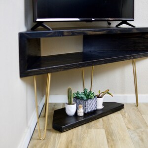 Puede incluir: Un mueble de TV de madera negra con patas doradas de horquilla. El mueble tiene un estante abierto y un estante inferior con peque&ntilde;as plantas en macetas y un cactus. El mueble est&aacute; en &aacute;ngulo y colocado en una esquina.