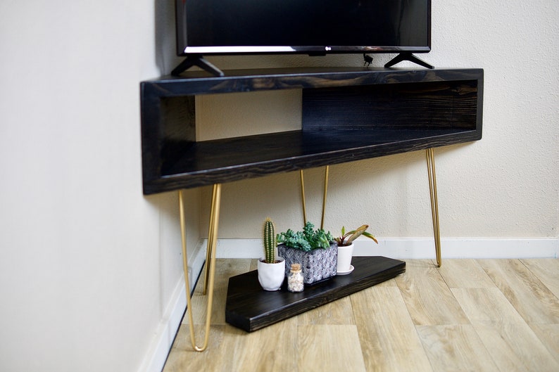 Puede incluir: Un mueble de TV de madera negra con patas doradas de horquilla. El mueble tiene un estante abierto y un estante inferior con peque&ntilde;as plantas en macetas y un cactus. El mueble est&aacute; en &aacute;ngulo y colocado en una esquina.