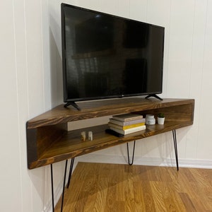 May include: A dark brown, corner TV stand with black hairpin legs. The stand has an open shelf with books, candles, and a small plant. A black flat-screen TV sits on top of the stand. The stand is placed in a corner against a white wall.