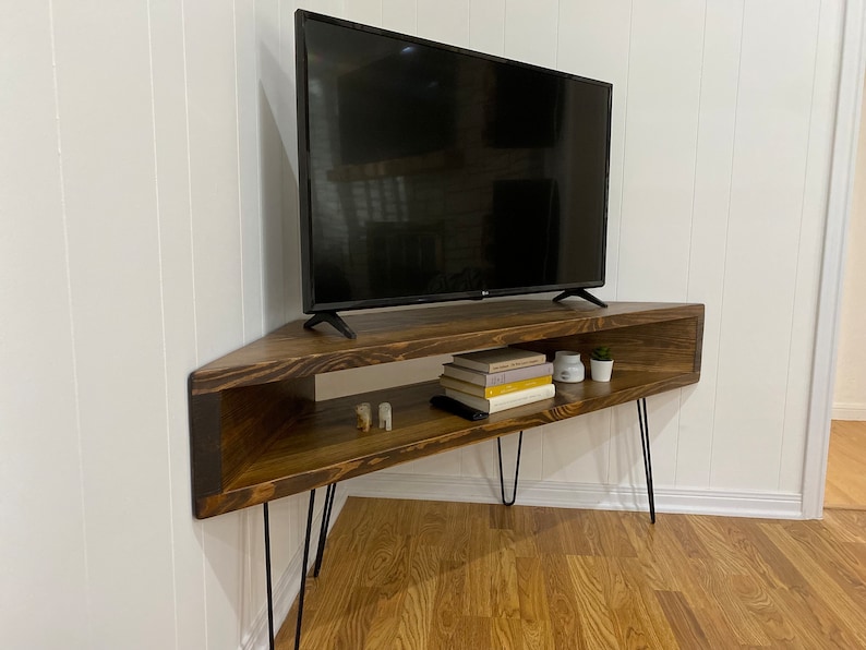 May include: A dark brown, corner TV stand with black hairpin legs. The stand has an open shelf with books, candles, and a small plant. A black flat-screen TV sits on top of the stand. The stand is placed in a corner against a white wall.