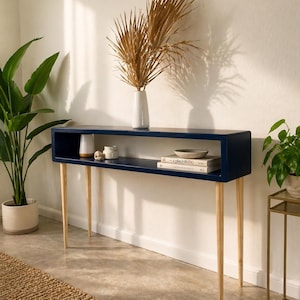 May include: A navy blue console table with a rectangular top and a lower shelf. The table has four slender, light-colored wooden legs. A white vase with dried palm fronds sits on top, along with decorative items and books. The table is 48 inches long.