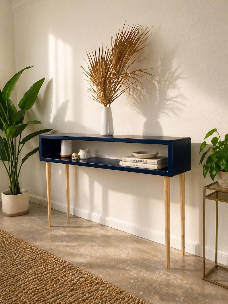 May include: A navy blue console table with a rectangular top and a lower shelf. The table has four slender, light-colored wooden legs. A white vase with dried palm fronds sits on top, along with decorative items and books. The table is 48 inches long.