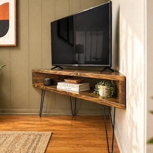May include: A dark wood corner TV stand with a black flat-screen TV. The stand has hairpin legs and a shelf with books, a small plant, and a candle. The wall behind the stand is olive green.