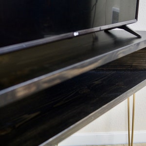 Puede incluir: Un mueble de TV de madera te&ntilde;ida oscura con un estante inferior y patas doradas de horquilla. El soporte sostiene un televisor negro. La veta de la madera es visible en la superficie del soporte.