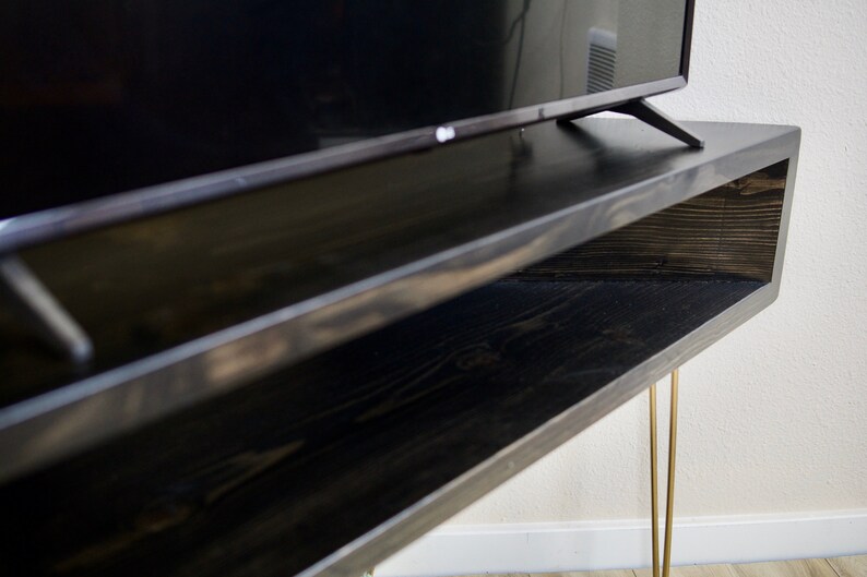 Puede incluir: Un mueble de TV de madera te&ntilde;ida oscura con un estante inferior y patas doradas de horquilla. El soporte sostiene un televisor negro. La veta de la madera es visible en la superficie del soporte.