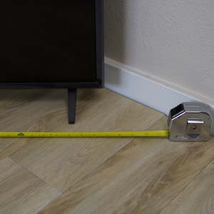 Puede incluir: Un televisor negro con una cinta m&eacute;trica extendida por el suelo. La cinta m&eacute;trica es amarilla con marcas negras y mide la distancia desde la esquina de la habitaci&oacute;n. El suelo es de color marr&oacute;n claro.