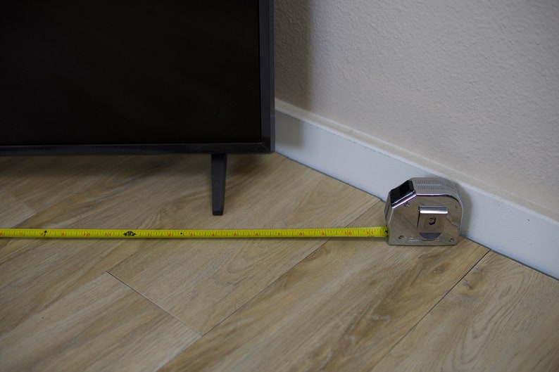 Puede incluir: Un televisor negro con una cinta m&eacute;trica extendida por el suelo. La cinta m&eacute;trica es amarilla con marcas negras y mide la distancia desde la esquina de la habitaci&oacute;n. El suelo es de color marr&oacute;n claro.