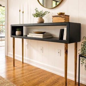 May include: A modern black console table with a rectangular top and open shelf. The table has four tapered wooden legs. Decorative items on the table include a small plant, books, and a wooden box. The table is placed against a white wall.