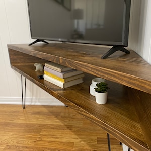 May include: A dark brown, corner-shaped TV stand with a flat-screen television on top. The stand has a lower shelf with books, decorative items, and a small plant. The stand is supported by black hairpin legs.
