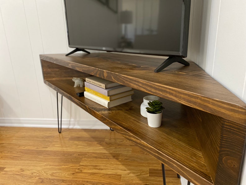 May include: A dark brown, corner-shaped TV stand with a flat-screen television on top. The stand has a lower shelf with books, decorative items, and a small plant. The stand is supported by black hairpin legs.