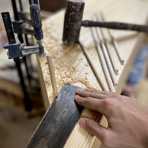 May include: A woodworking scene with a hand saw cutting a wooden dowel. A mallet, clamps, and several dowels are also visible on the wooden surface, along with wood shavings. The tools suggest a carpentry project in progress.
